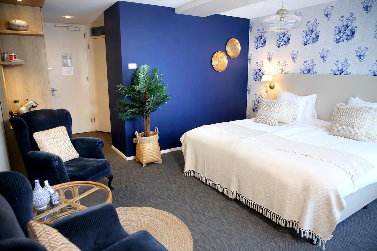 A hotel room with a large bed, a dark blue accent wall, a potted plant, and a wicker side table.