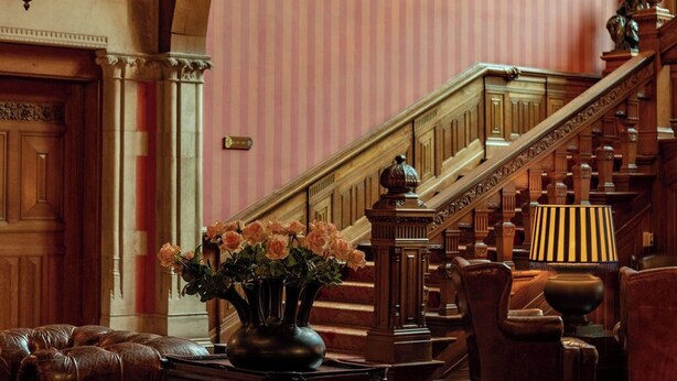 A grand lobby with a wooden staircase, leather seating, a chandelier, and a painting on the wall.