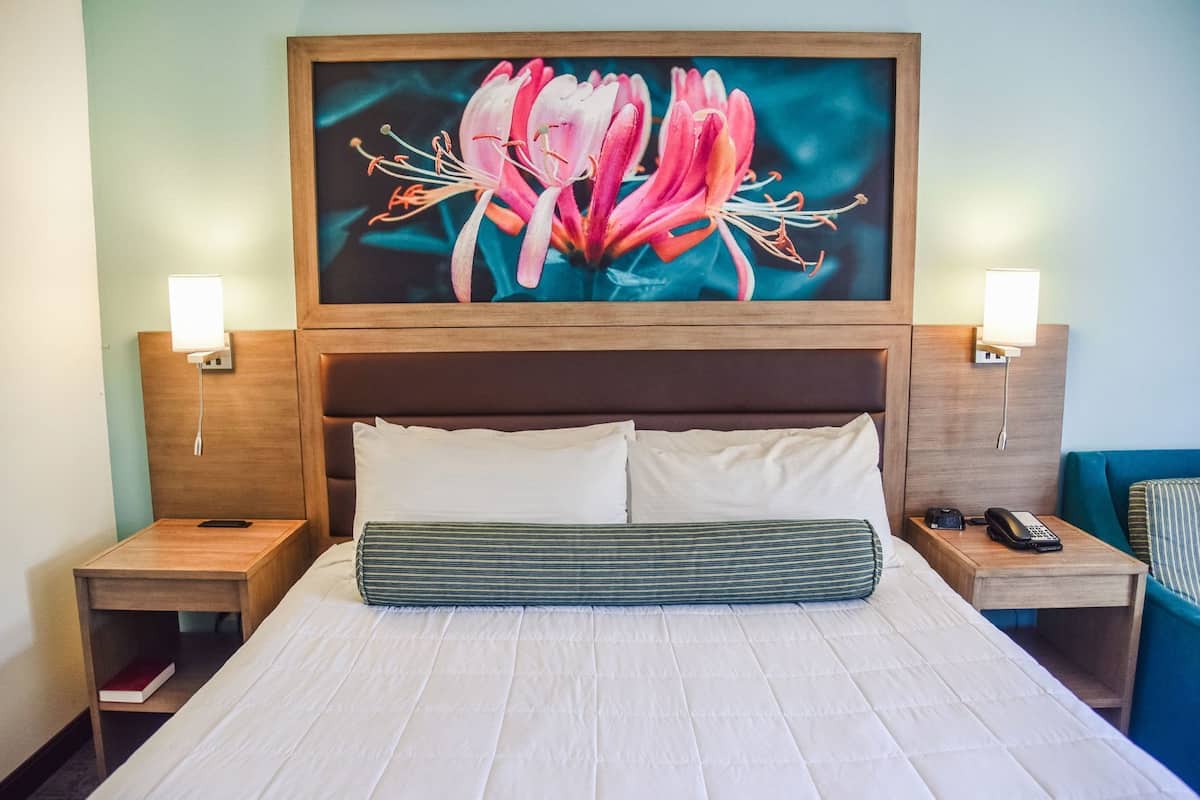 A hotel room with a large bed, two bedside tables, a nightstand, a painting above the bed, and a lamp on each bedside table.