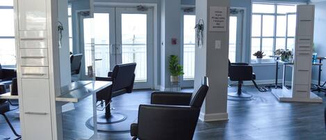 Een moderne salon met meerdere zwarte kappersstoelen, een wit receptiebalie en grote ramen die natuurlijk licht binnenlaten.