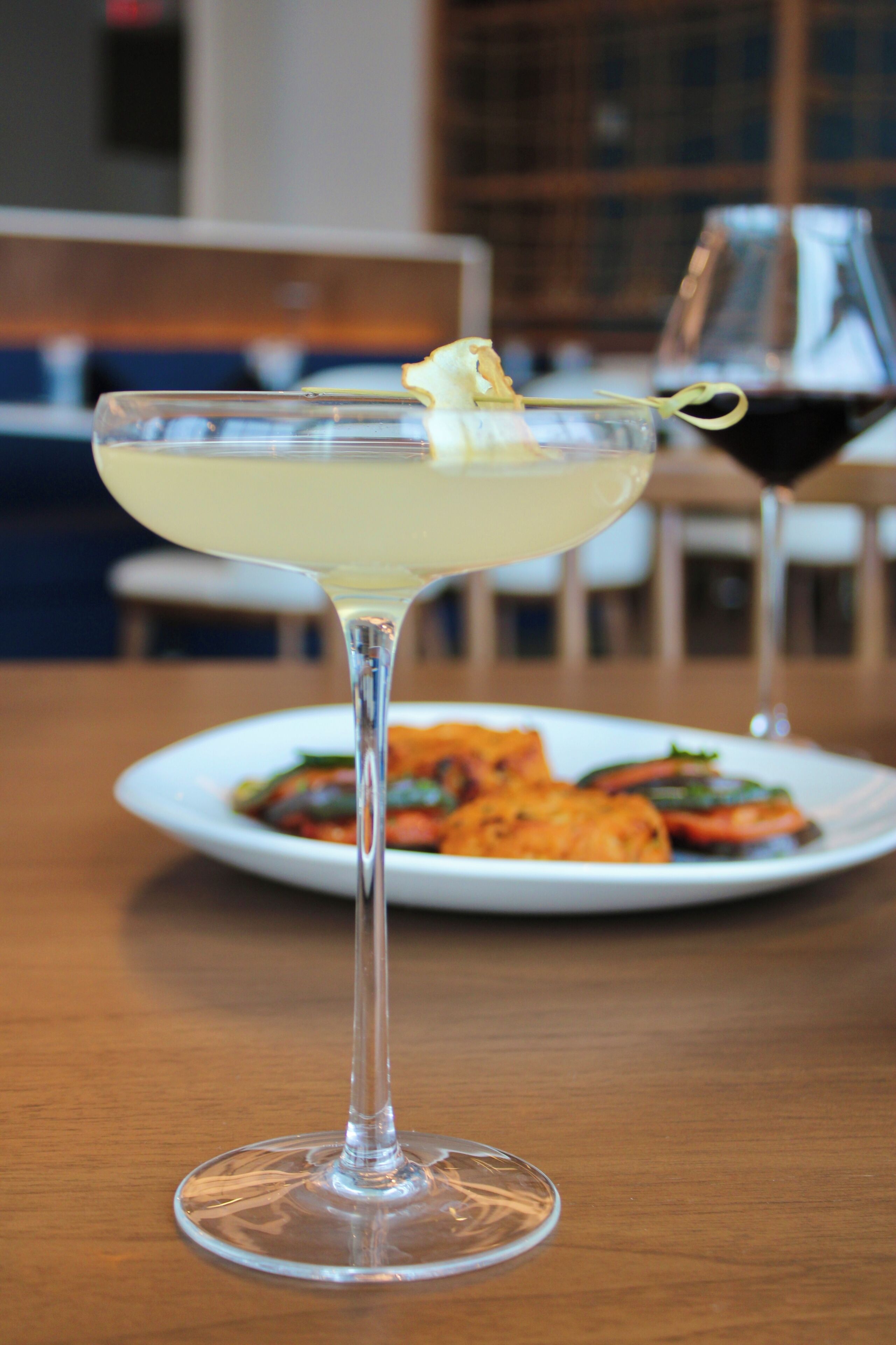 A cocktail glass with a lemon garnish on a wooden table, with a plate of food and a wine glass in the background.