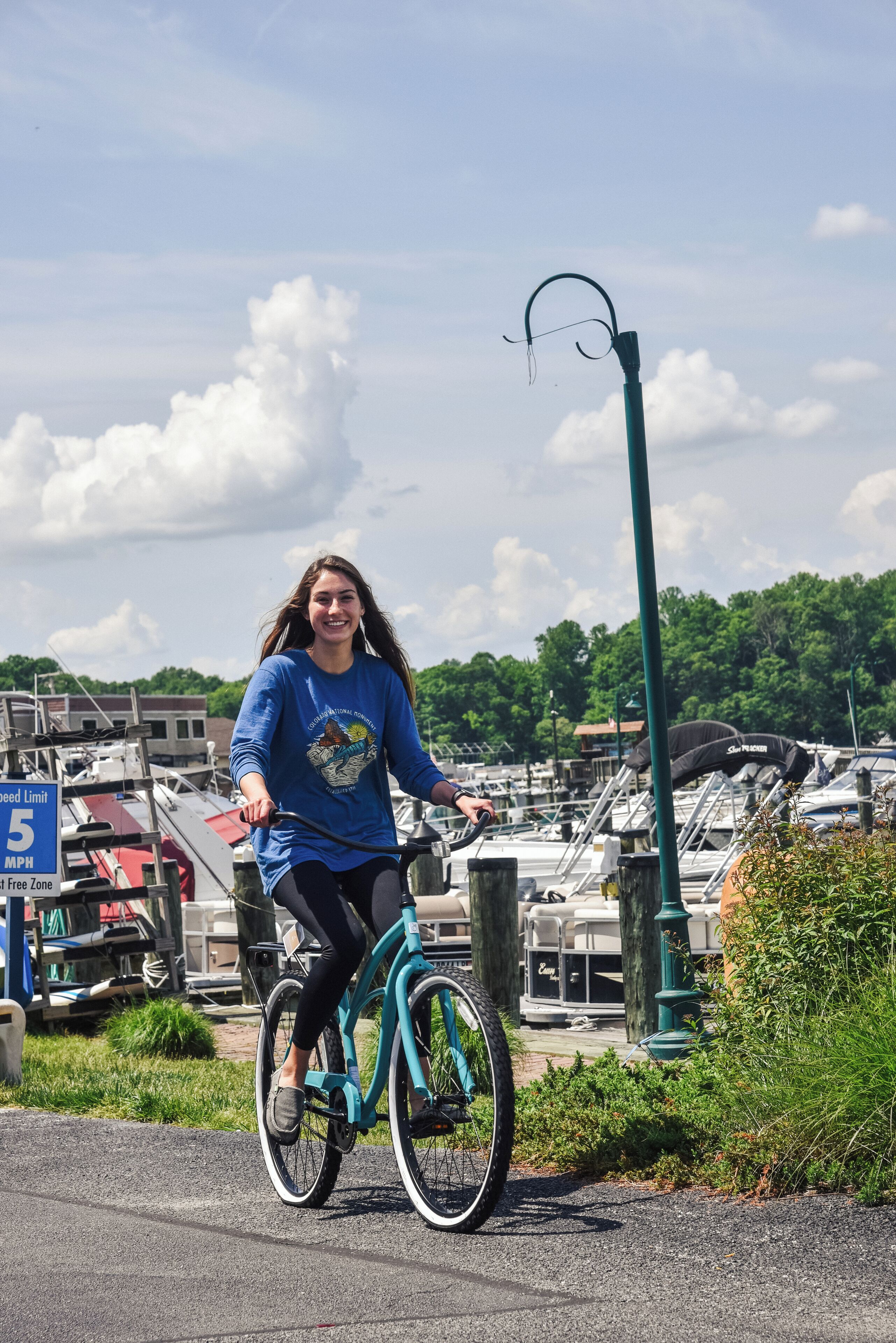 Eine Person fährt auf einem Radweg, im Hintergrund liegen Boote im Hafen.