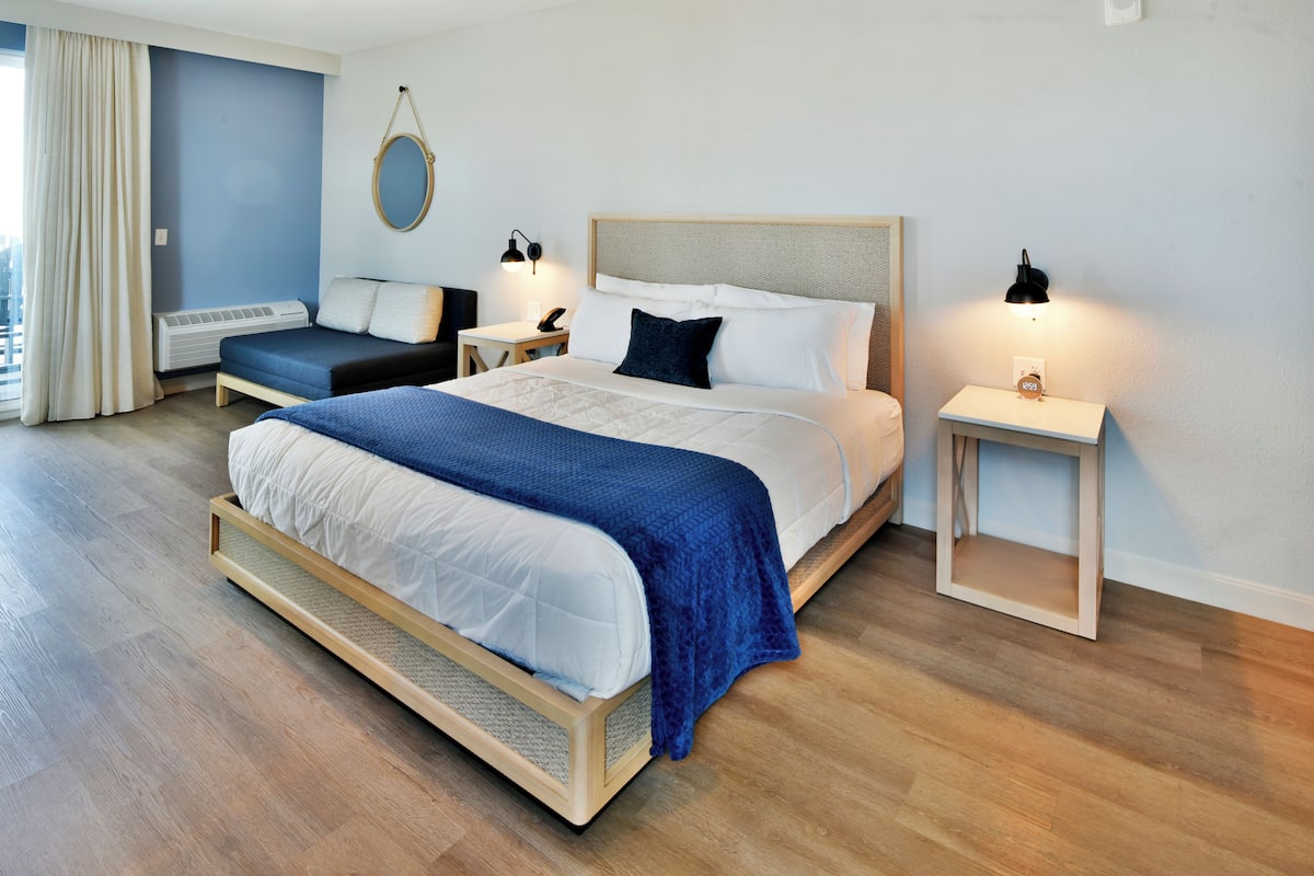 A modern hotel room with a large bed, two bedside tables, a wall-mounted mirror, and a view of the city.