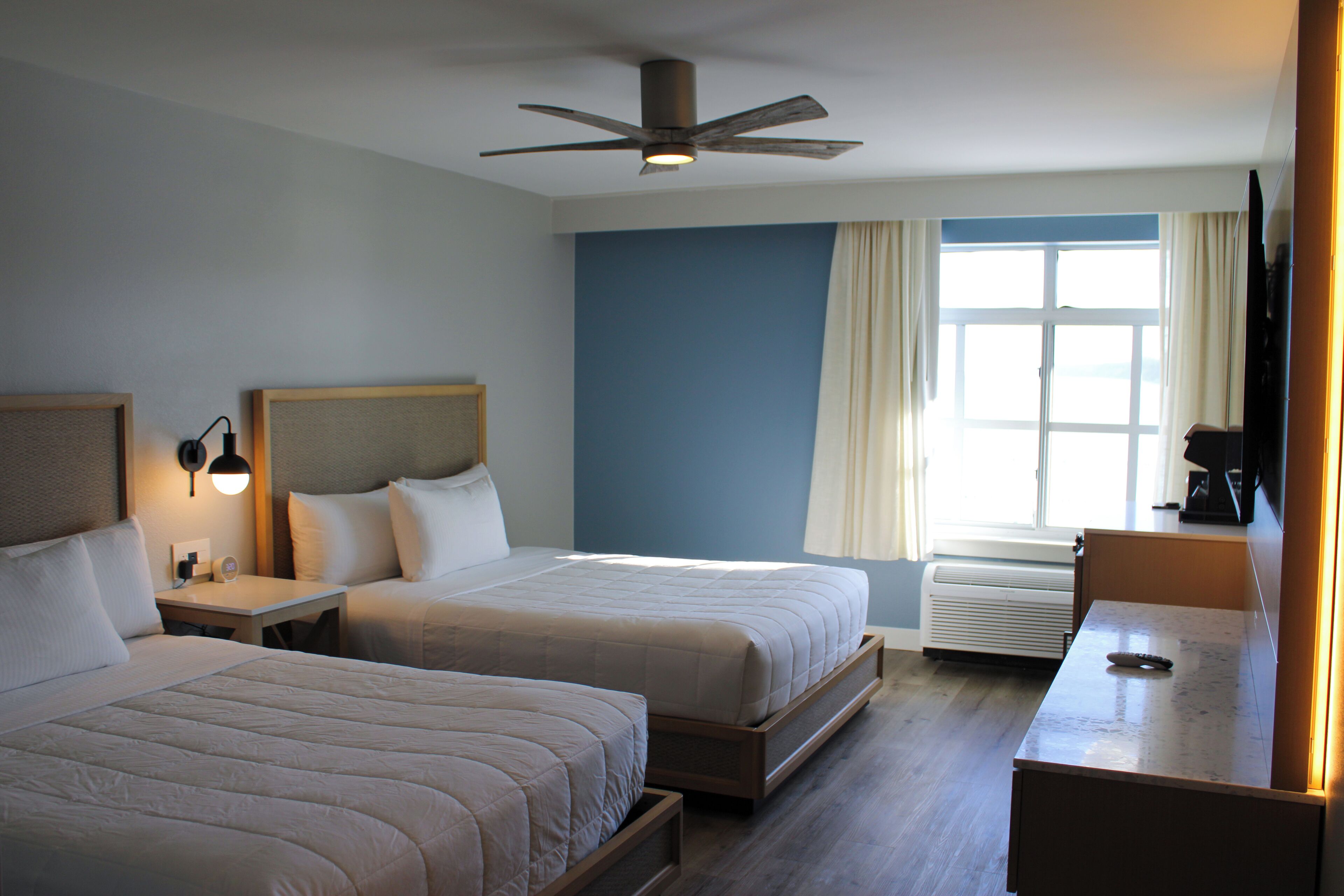 Habitación de hotel con dos camas, ventilador de techo, ventana con cortinas, escritorio y cafetera.