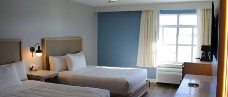 Habitación de hotel con dos camas, ventilador de techo, ventana con cortinas, escritorio y cafetera.
