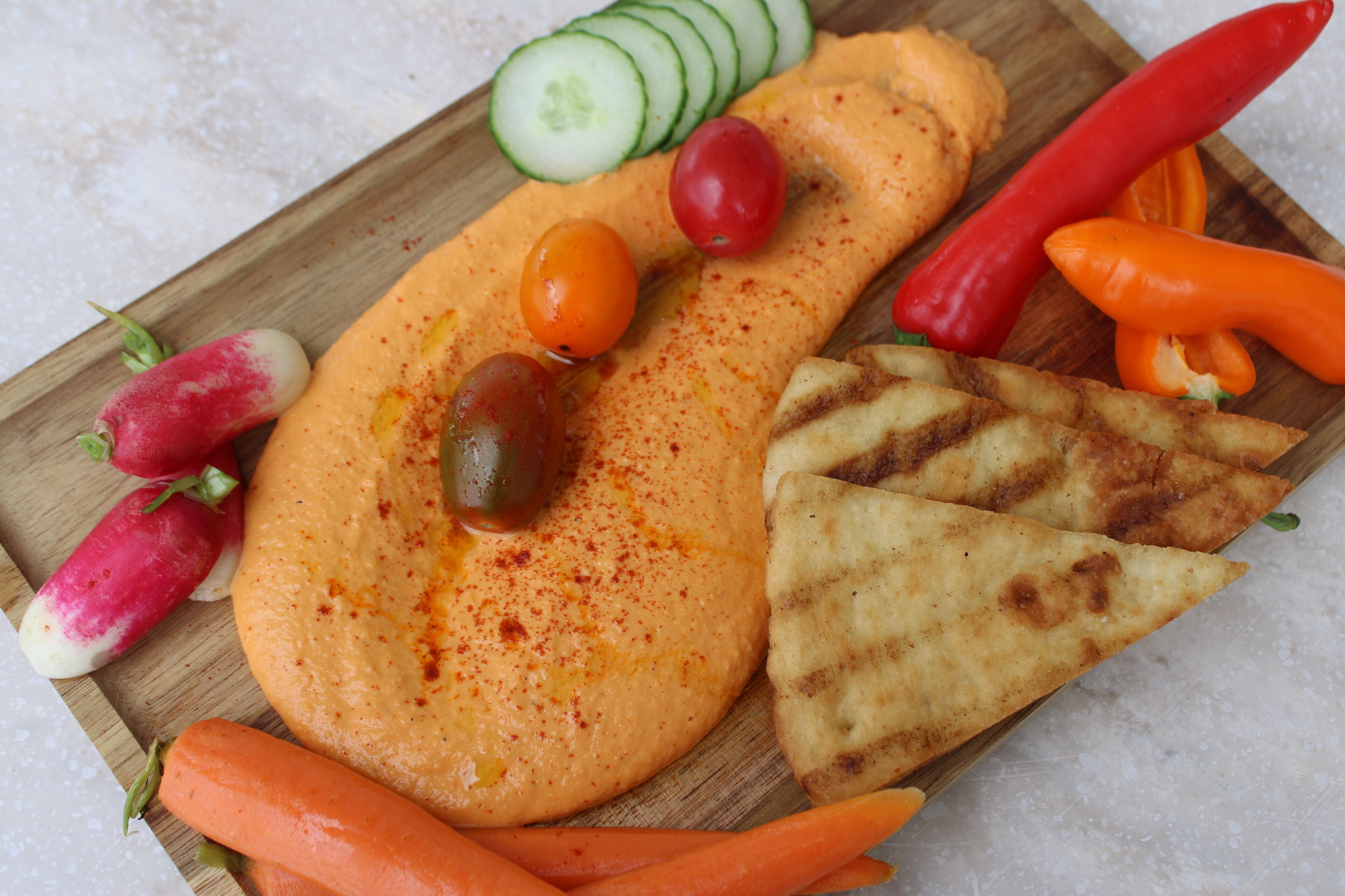 En träbricka med hummus, skivade gurkor, körsbärstomater, rädisor, morötter, grillat pitabröd samt röda och gula paprikor.