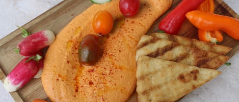 En träbricka med hummus, skivade gurkor, körsbärstomater, rädisor, morötter, grillat pitabröd samt röda och gula paprikor.