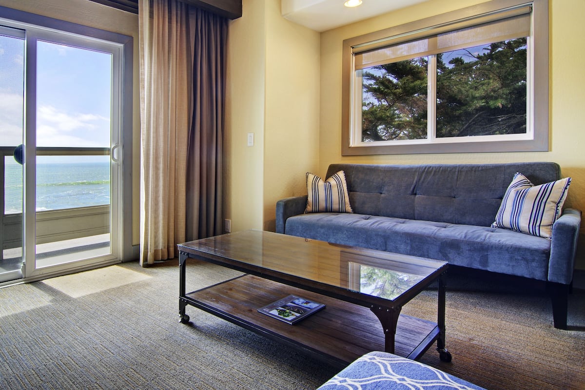 A living room with a sofa, a coffee table, and a view of the ocean through large windows.