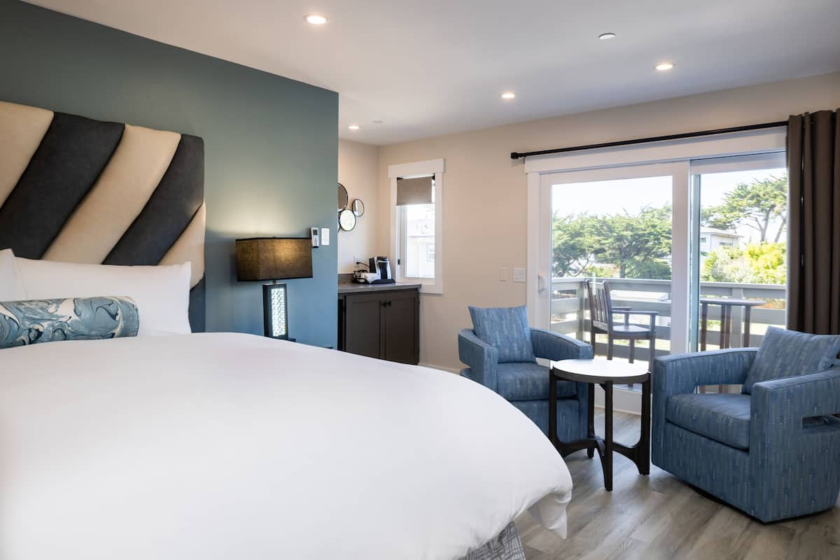 A modern hotel room with a large bed, a desk, two armchairs, and a balcony with outdoor seating.