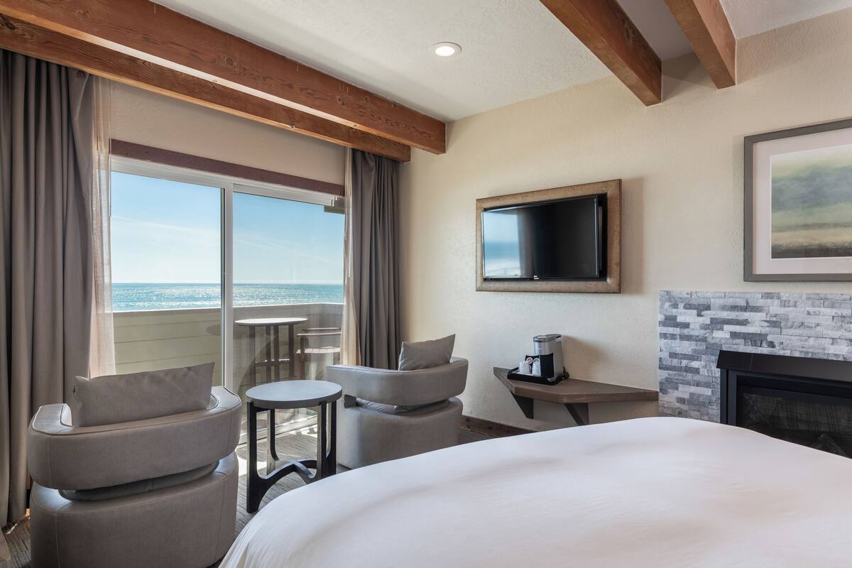 A hotel room with a bed, two armchairs, a small table, a TV, and a fireplace.