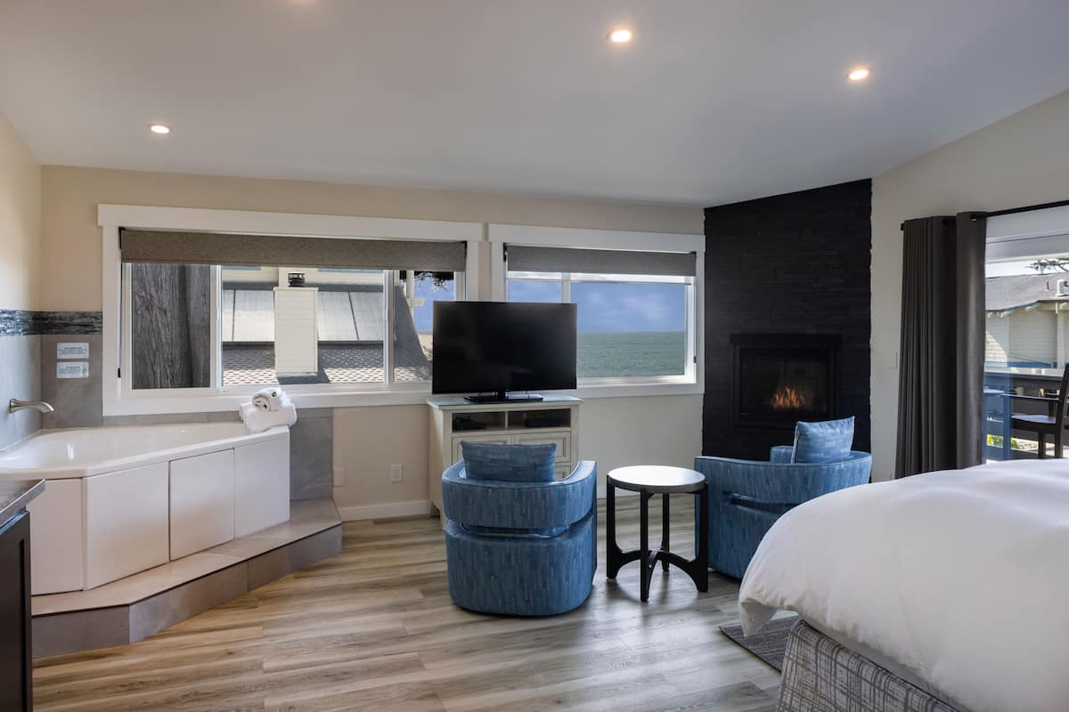 A modern hotel room with a large bed, a TV, a fireplace, a bathtub, and a view of the sea.