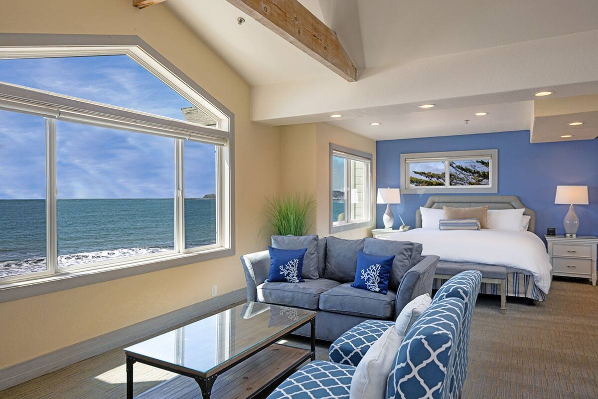 A bedroom with a large window overlooking the sea, a bed with blue bedding, a sofa with blue cushions, and a bedside table.