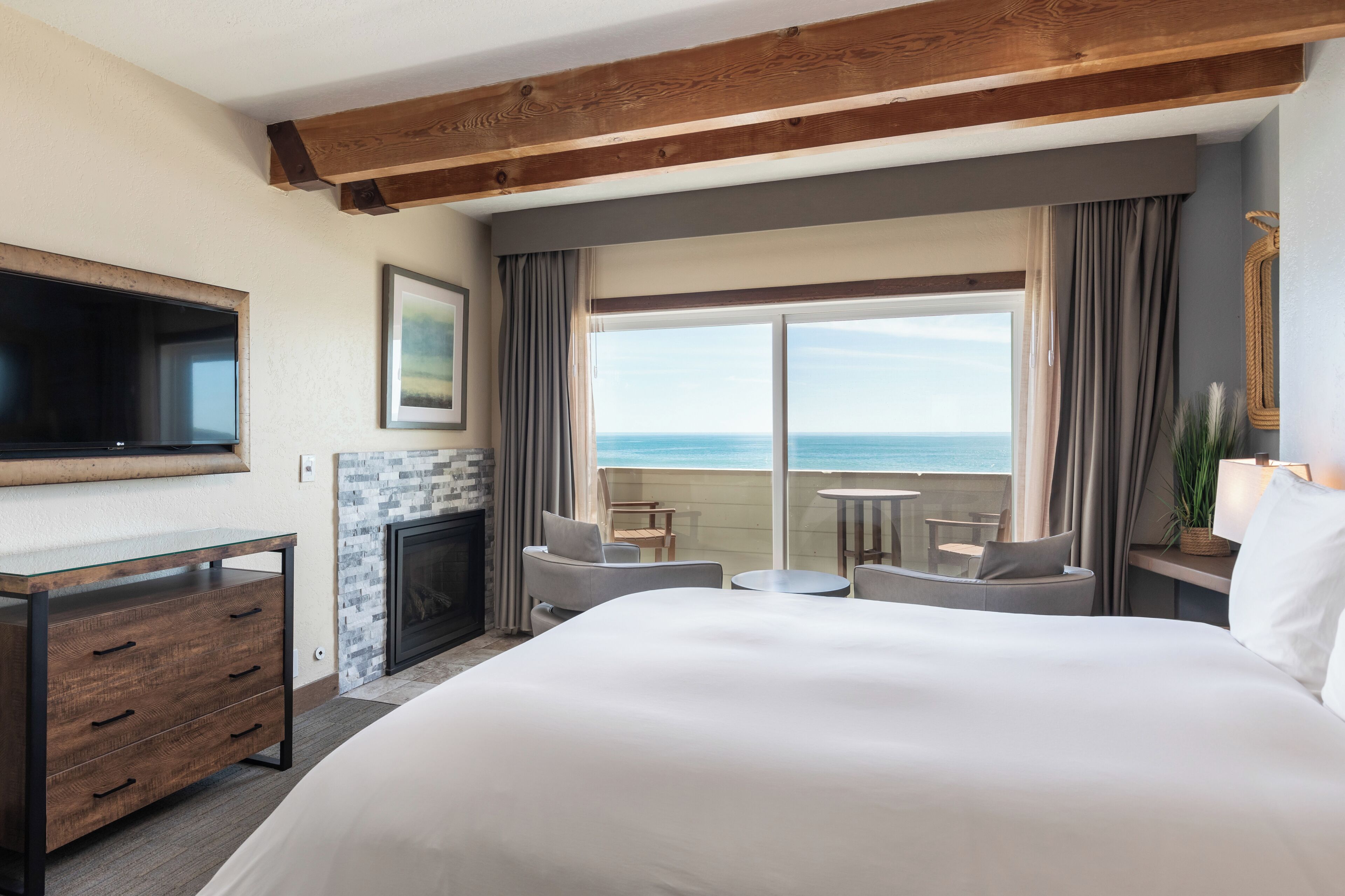 Quarto de hotel com uma cama grande, televisão na parede, lareira, varanda com vista para o mar e uma pequena mesa com cadeiras.
