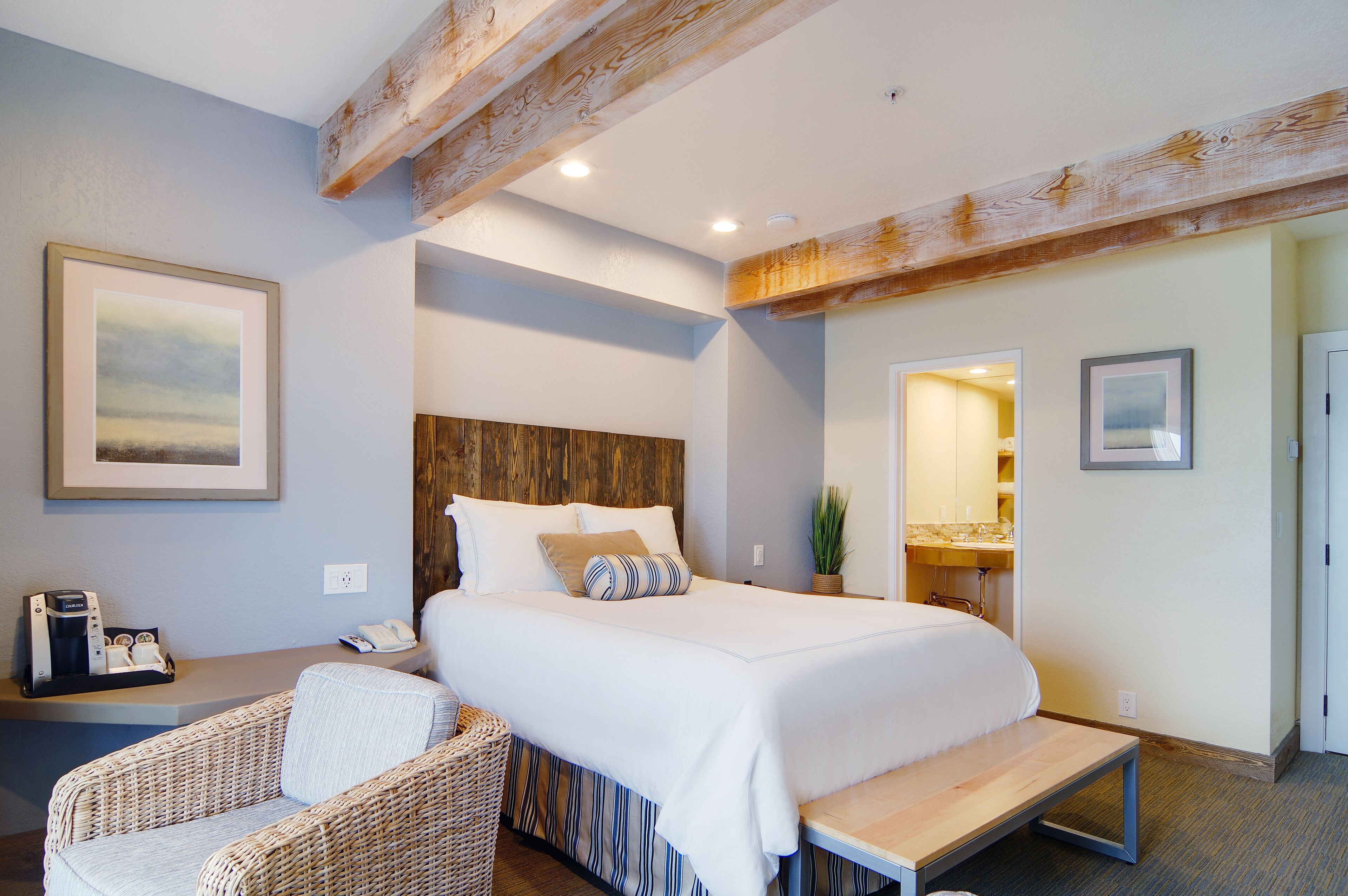 Ein Hotelzimmer mit einem großen Bett, einem Holzkopfende, einem Korbstuhl, einem Nachttisch mit Telefon und einer Kaffeemaschine sowie einem durchgegangenen Tür ersichtlichen Badezimmer.