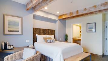 A hotel room with a large bed, a wooden headboard, a wicker chair, a bedside table with a phone and a coffee maker, and a bathroom visible through an open door.