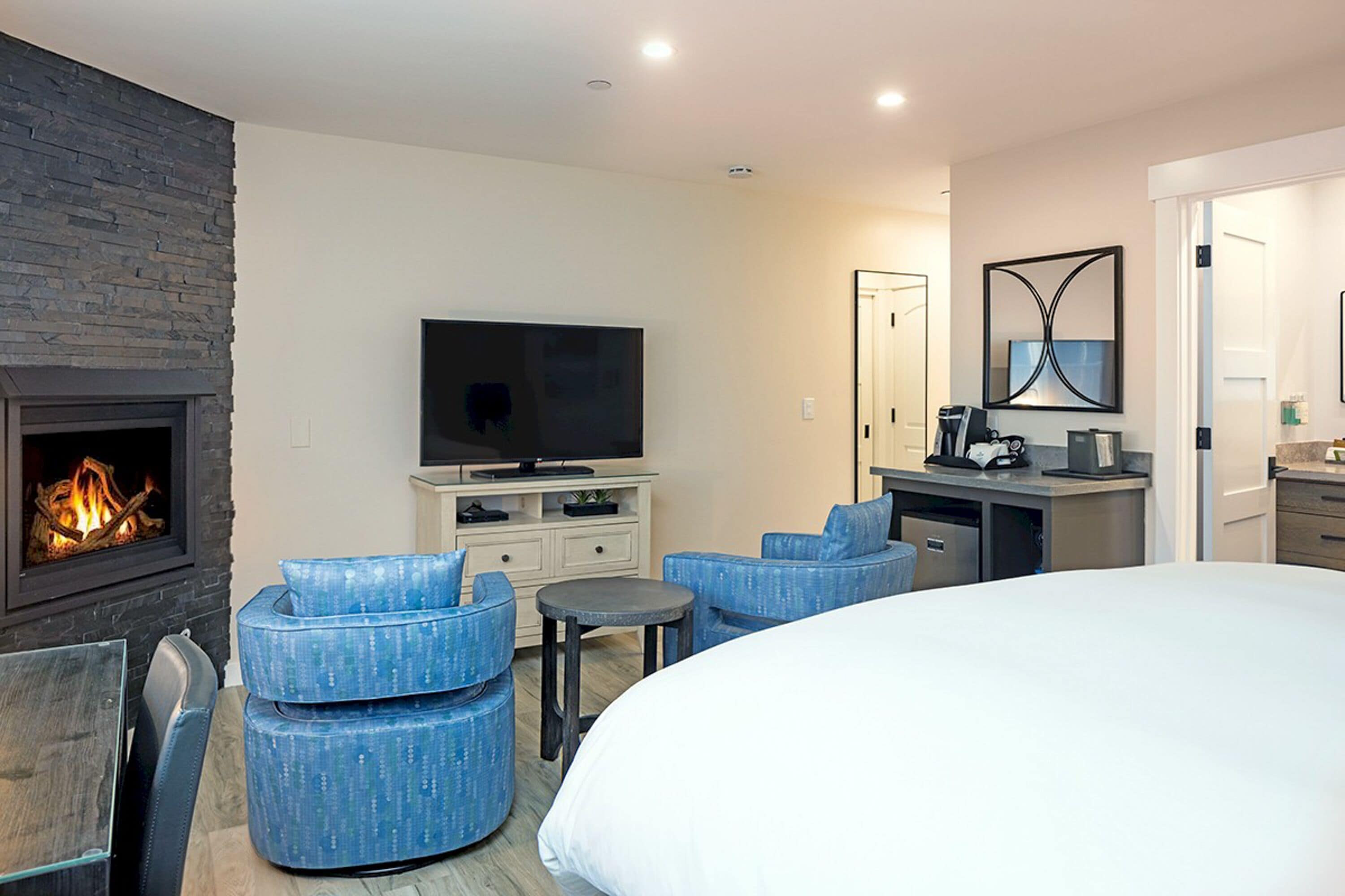 A modern hotel room with a fireplace, a flat-screen TV, a large bed, and a seating area with blue upholstered chairs.