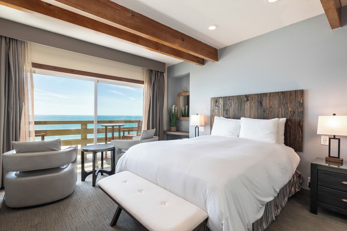 A hotel room with a large bed, a bench, a desk, and a view of the ocean.