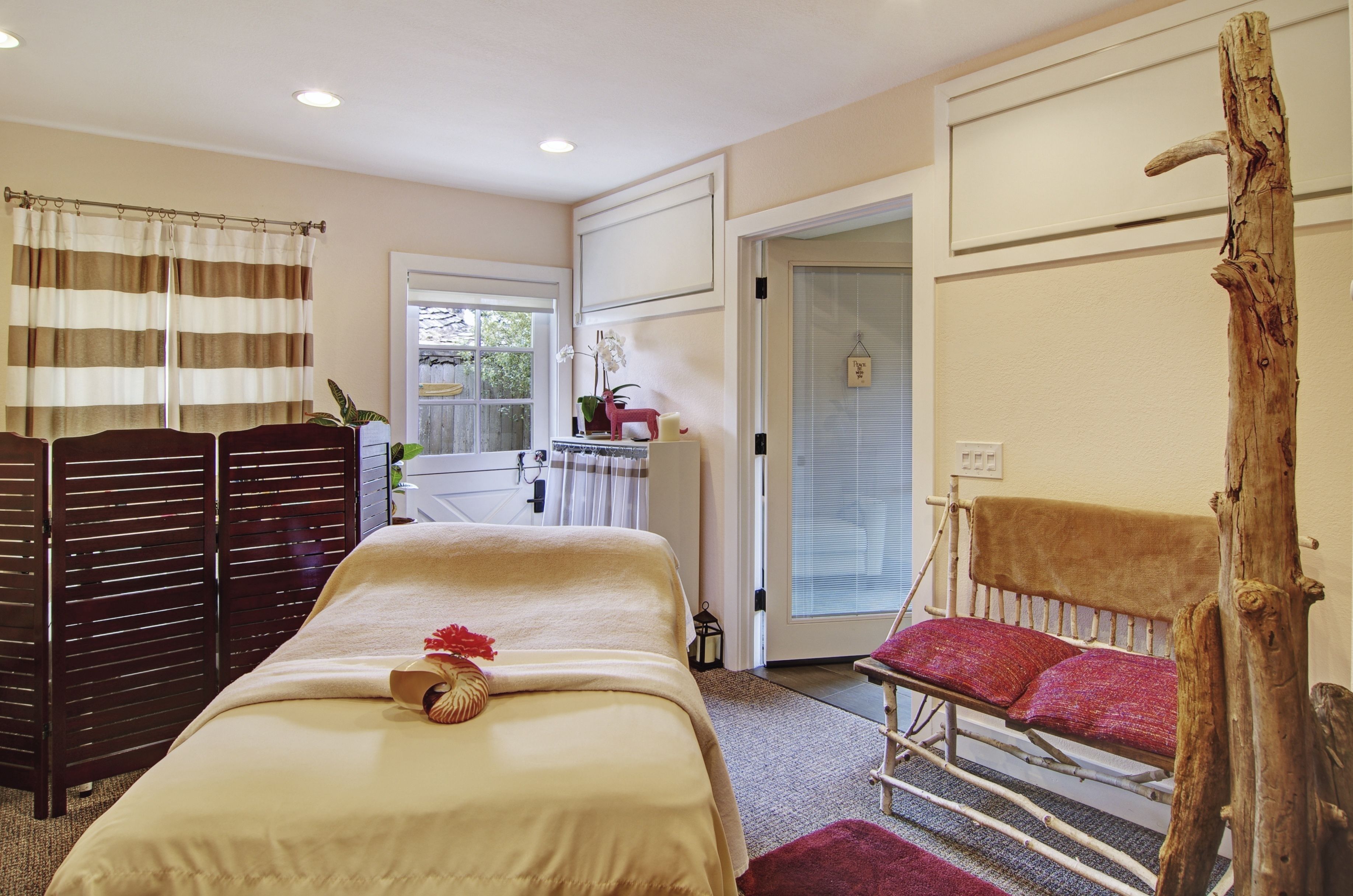 Un dormitorio con cama, una silla de madera, un banco, una cortina y una ventana.