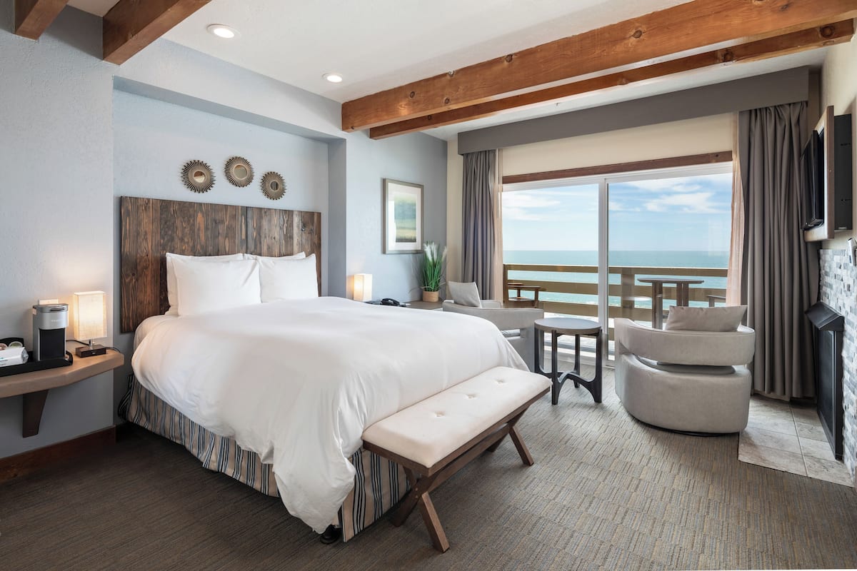 A hotel room with a large bed, a bench, a desk with a lamp, a chair, and a view of the sea.