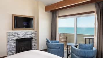 Quarto de hotel com lareira, televisão na parede, varanda com vista para o mar e dois cadeiras e uma mesa.