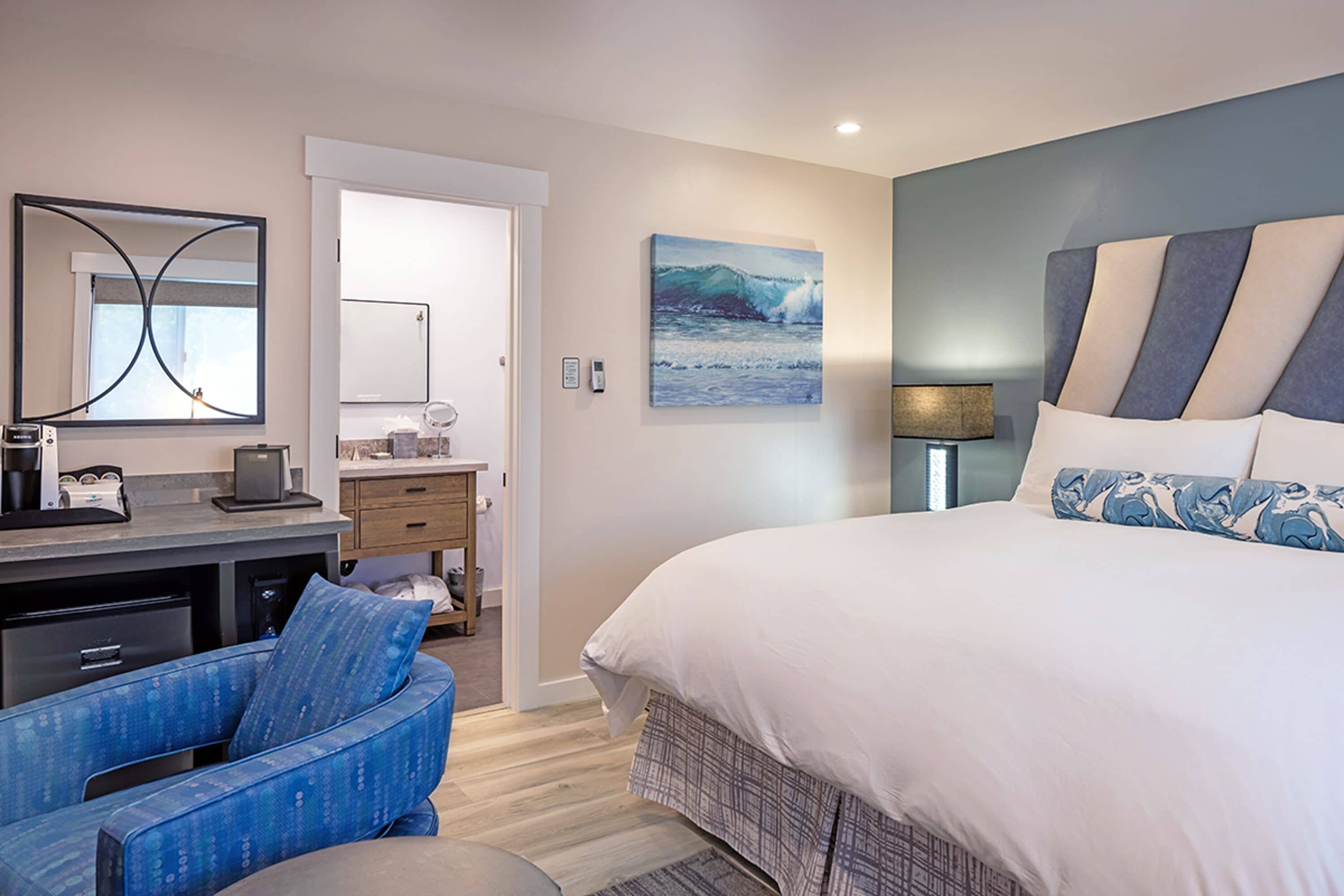 A bedroom with a large bed, a desk with a chair, a bathroom with a mirror, and a painting on the wall.