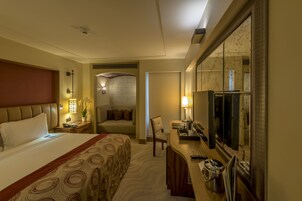 A hotel room with a large bed, a desk with a TV, a chair, and a small table with a lamp.