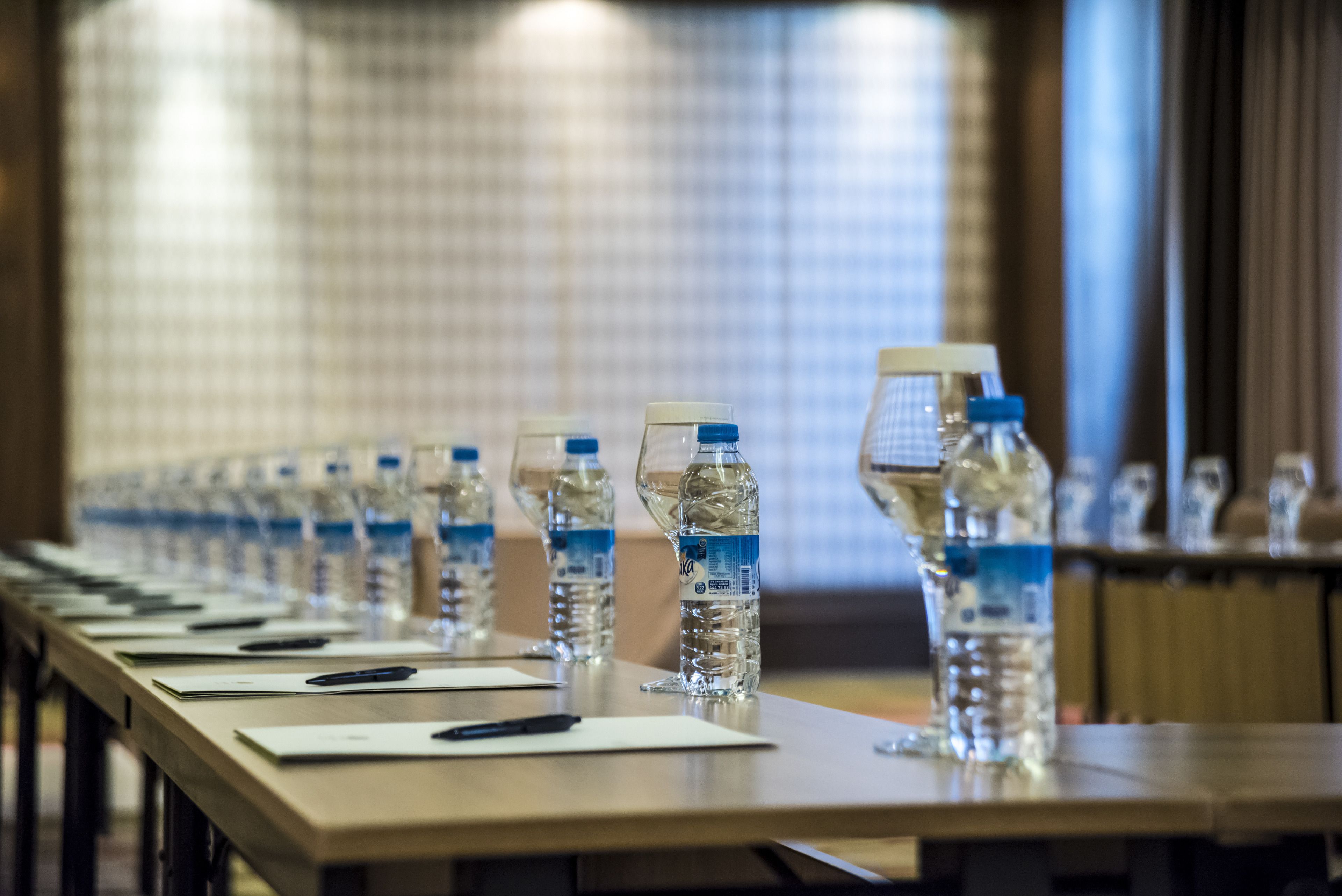 Una mesa de conferencias con botellas de agua y bolígrafos, frente a una pared de azulejos.