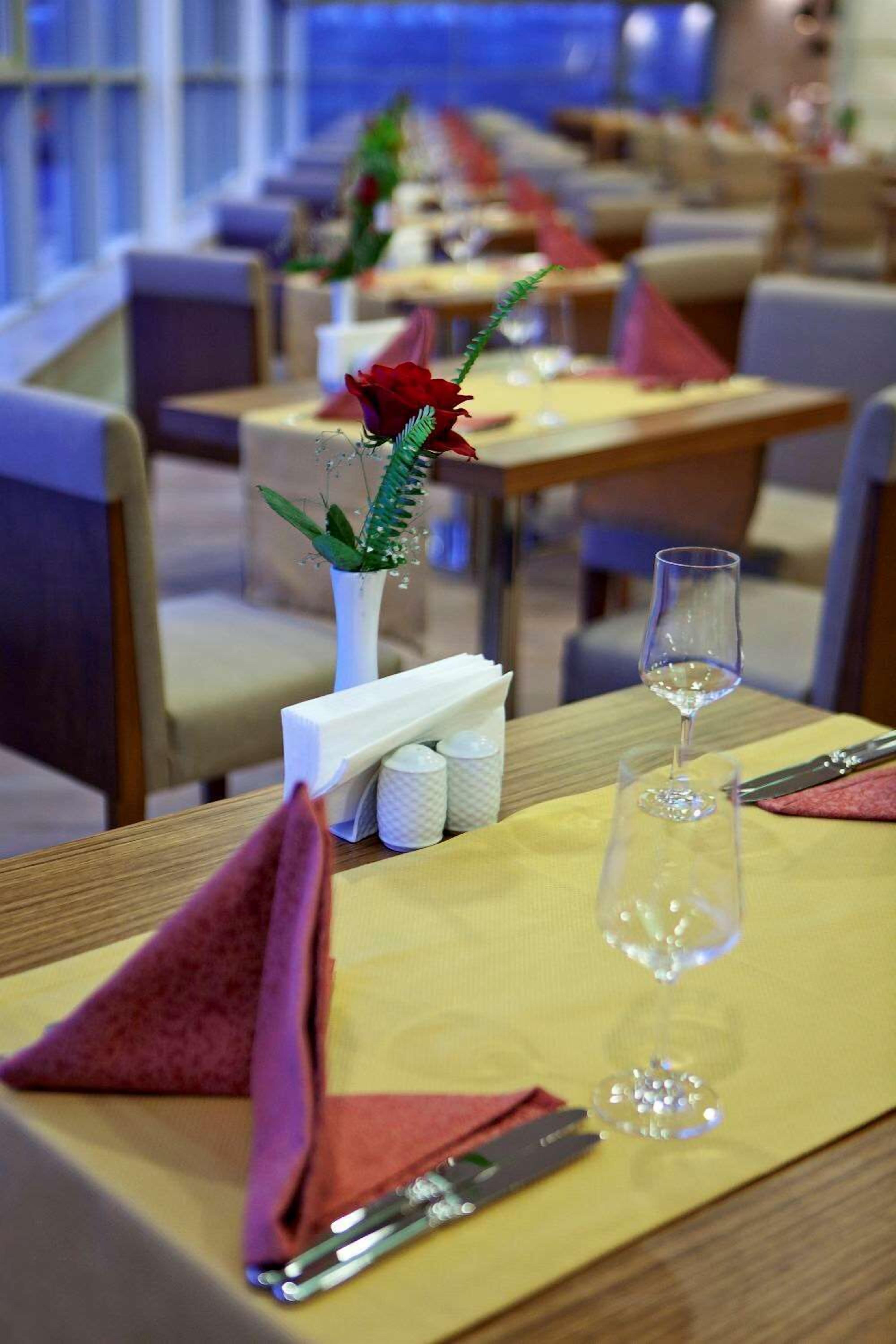 Ambiente de restaurante com mesas preparadas para refeições, destacando um vaso com uma rosa vermelha e uma taça de vinho.
