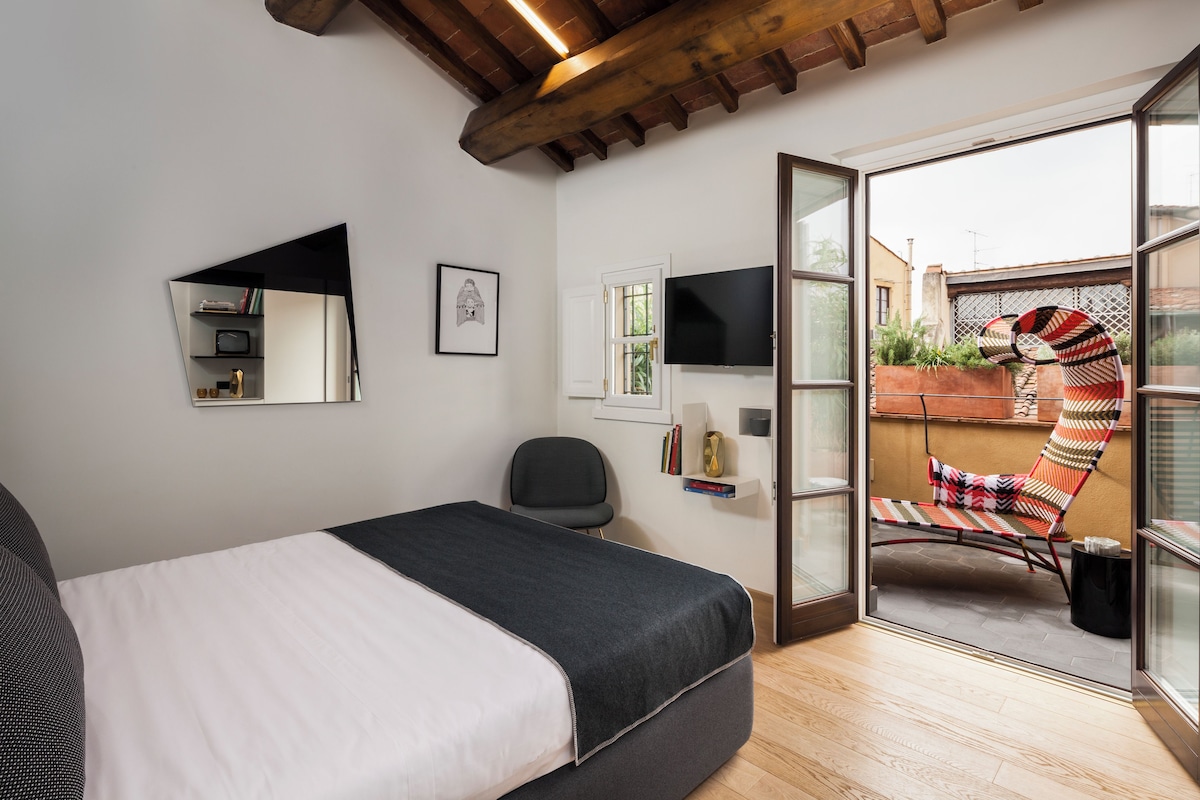 A bedroom with a bed, a chair, a TV, and a balcony with a chair and plants.