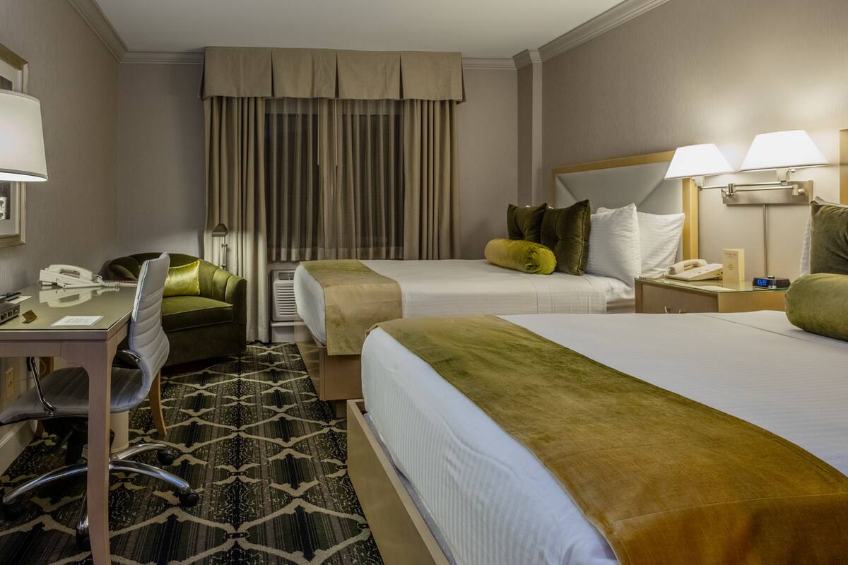 Quarto de hotel com duas camas, uma escrivaninha com telefone, uma cadeira e janela com cortinas.