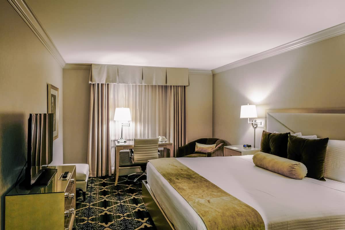 Quarto de hotel com uma cama grande, uma escrivaninha com cadeira, televisão e luminária.