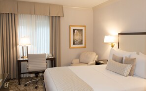 Habitación de hotel con cama, dos sillas, un escritorio con lámpara, teléfono y un cuadro en la pared.