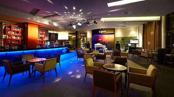Zona de bar con un mural de paisaje urbano, diferentes distribuciones de asientos y un escenario con equipo de iluminación.