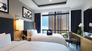 A hotel room with two beds, a desk with a chair, a large window with a city view, and artwork on the walls.