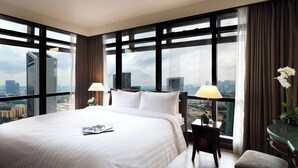 A hotel room with a large bed, two bedside tables, a chair, and a view of the cityscape through large windows.