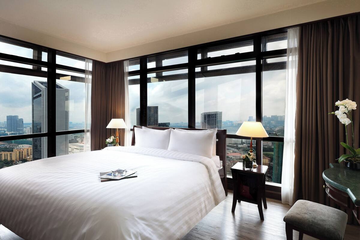 Une chambre d’hôtel avec un grand lit, deux tables de chevet, une chaise et une vue sur le paysage urbain grâce à de grandes fenêtres.
