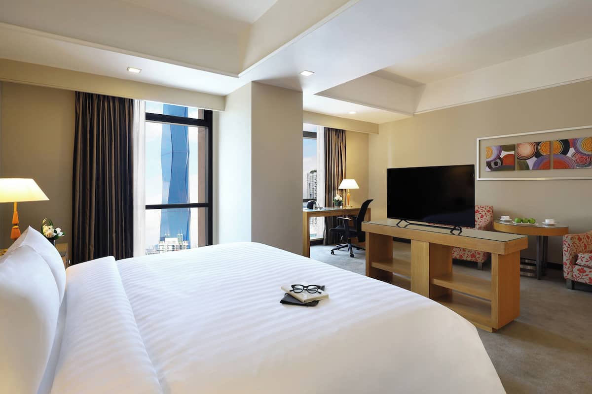 Une chambre d’hôtel avec un grand lit, un bureau doté d’un téléviseur à écran plat, une chaise, une petite table avec une tasse et des pommes vertes, et une fenêtre avec des rideaux.