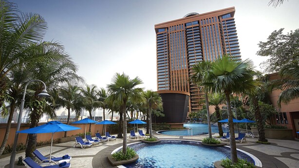 A hotel pool area with lounge chairs and umbrellas, surrounded by palm trees and a tall building in the background.