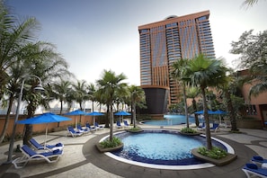 Zona de piscina con sillones de playa y sombrillas, rodeada de palmeras y un edificio alto al fondo.