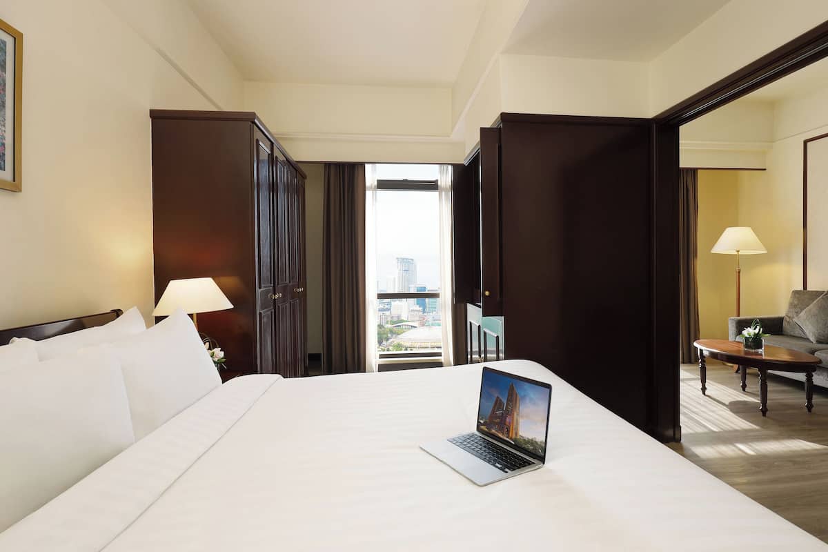 Une chambre d’hôtel avec un grand lit, un ordinateur portable posé sur le lit, une vue sur la ville par la fenêtre, un canapé et une lampe.