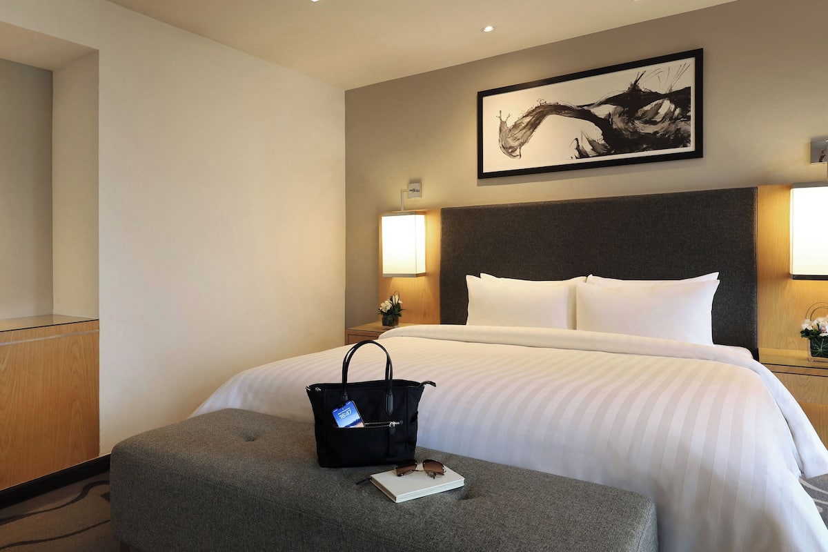 Une chambre d’hôtel avec un grand lit, une tête de lit, une table de chevet avec une lampe, une œuvre d’art encadrée au mur, et une petite table sur laquelle se trouvent un sac à main et un livre.