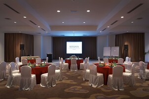 A conference room with a large screen displaying "BERJAYA" and a presentation. Tables are set with red tablecloths and white chairs.
