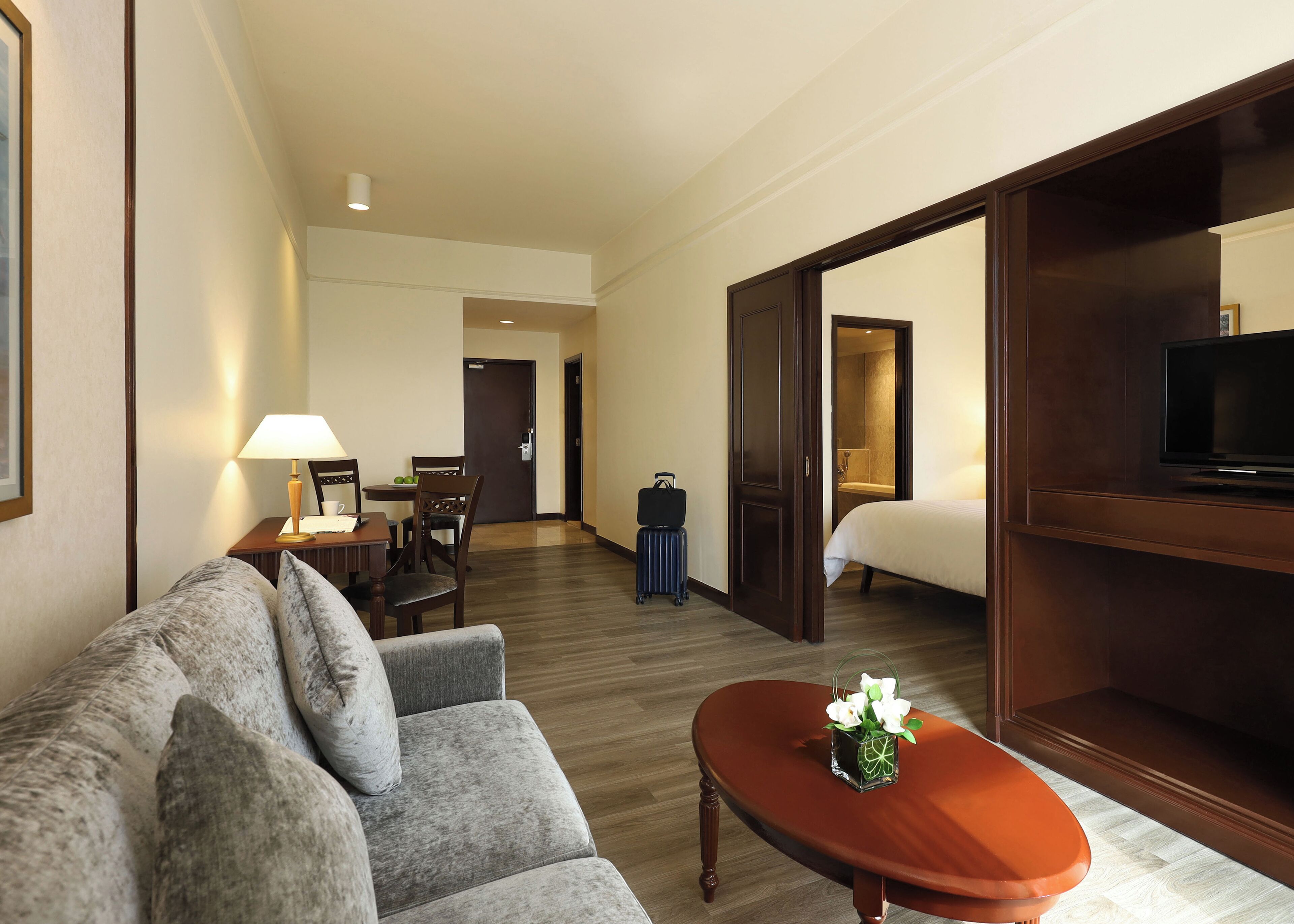 A hotel room with a sofa, a small table with a vase, a bed, a TV, and a bathroom visible through an open door.