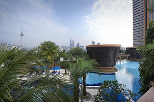 A swimming pool with palm trees and lounge chairs, surrounded by modern buildings and a clear sky.