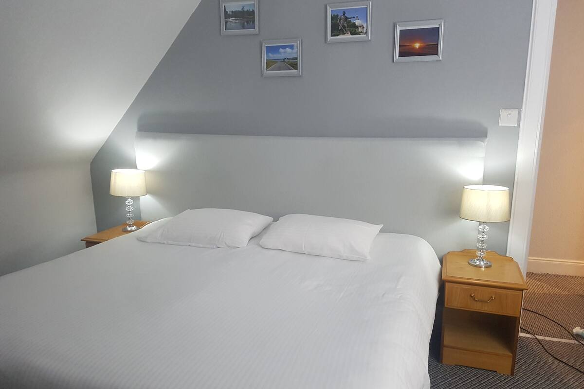 A neatly made bed with white linens in a room with a sloped ceiling. There are two bedside tables with lamps, and three framed pictures on the wall above the bed.
