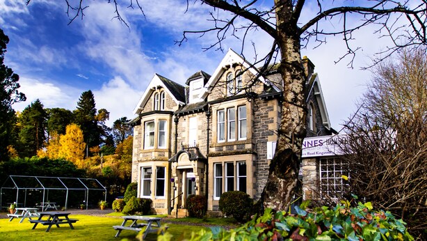 A historic stone building with a large tree in the foreground, a tennis court, and picnic tables. - McInnes House rooms with breakfast (Kingussie)