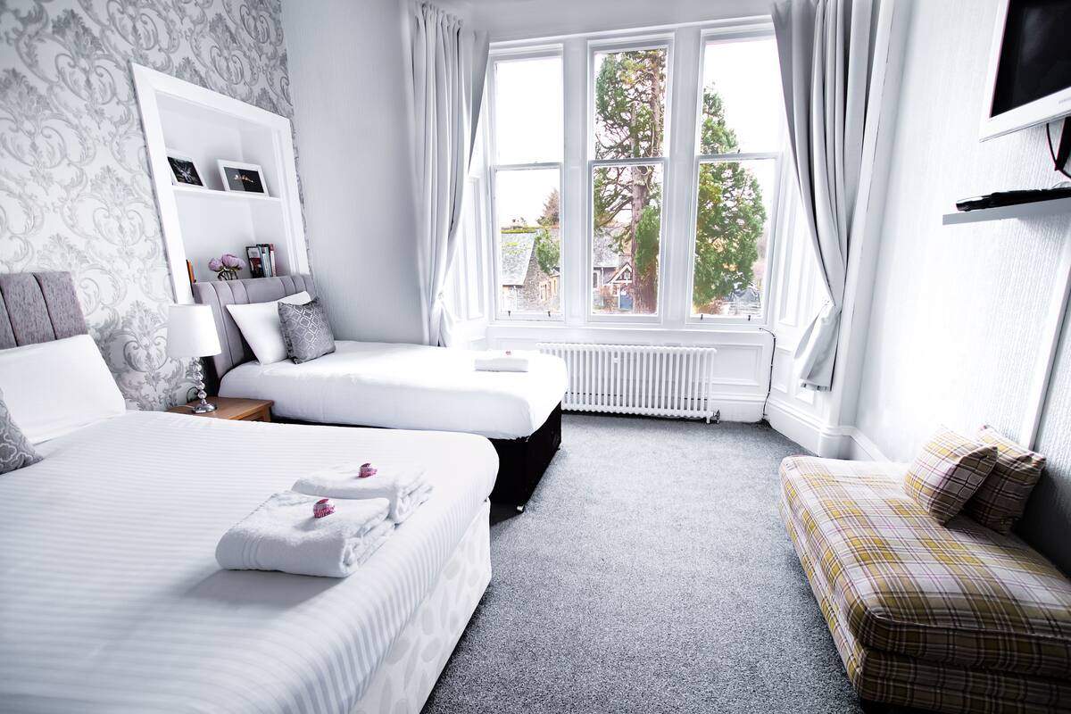 A hotel room with two beds, a sofa, a TV, and a window with a view of trees and houses.