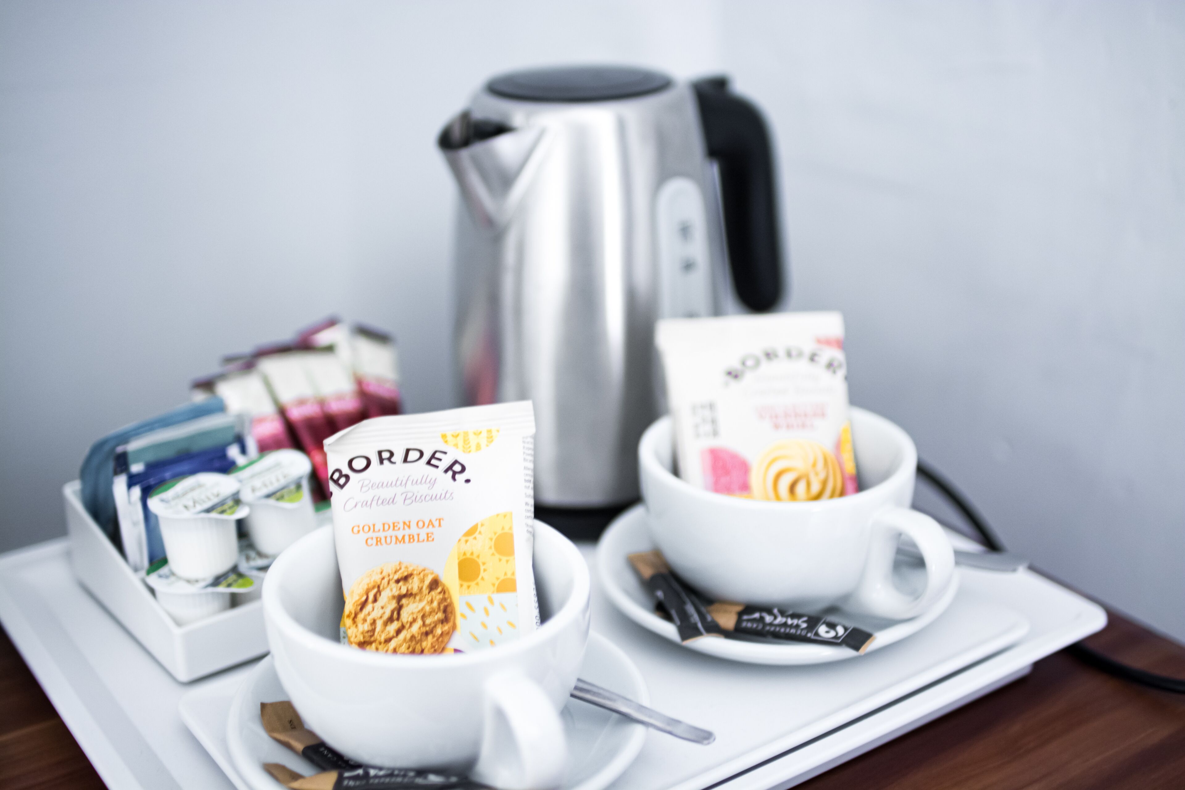 A breakfast setup with two cups of coffee, a kettle, and a tray with food items and a book.