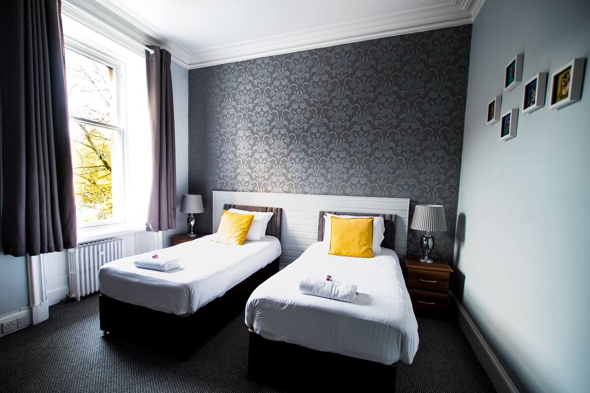 A hotel room with two beds, a window with curtains, a radiator, and a wall with framed pictures.