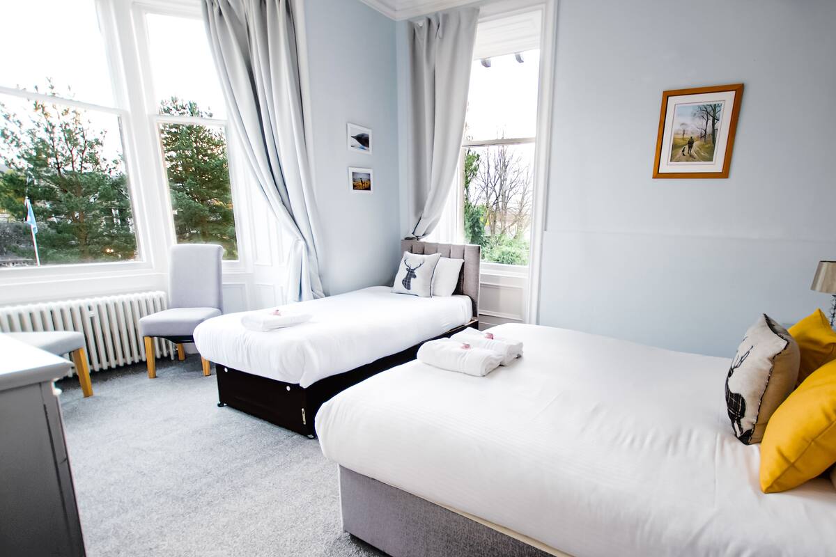 A hotel room with two beds, a desk, and a chair. There are curtains, a radiator, and a window with a view of trees.